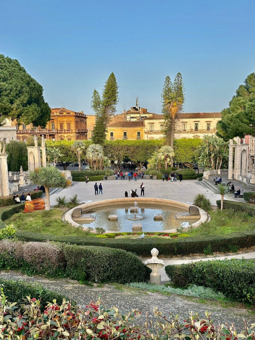 La vasca circolare del giardino con il getto d'acqua, il piazzale e i palazzi di Catania sullo sfondo